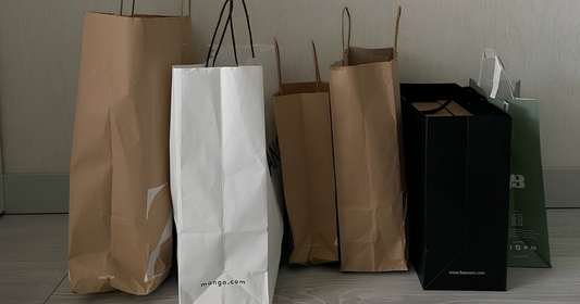 A group of shopping bags standing upright against a plain indoor wall, including several brown paper bags, one white paper bag, one black bag, and one green bag.
