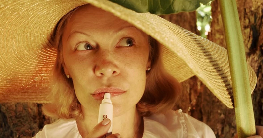 Pamela Anderson applying lipstick outdoors, wearing a wide-brimmed hat.
