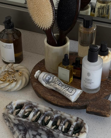 A bathroom vanity scene with several hair care and grooming products arranged on a countertop and wooden tray, including a tube labeled Maxval, bottles, brushes, and a marble hair comb in the foreground.