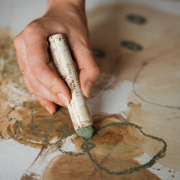 Hand holding a paintbrush with a textured handle on a wooden surface with artistic designs.